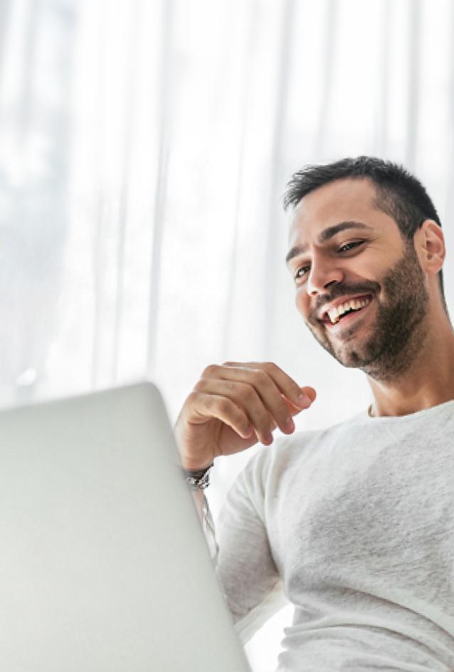 Man sitting in front of sheer blinds laughs while looking at laptop screen.