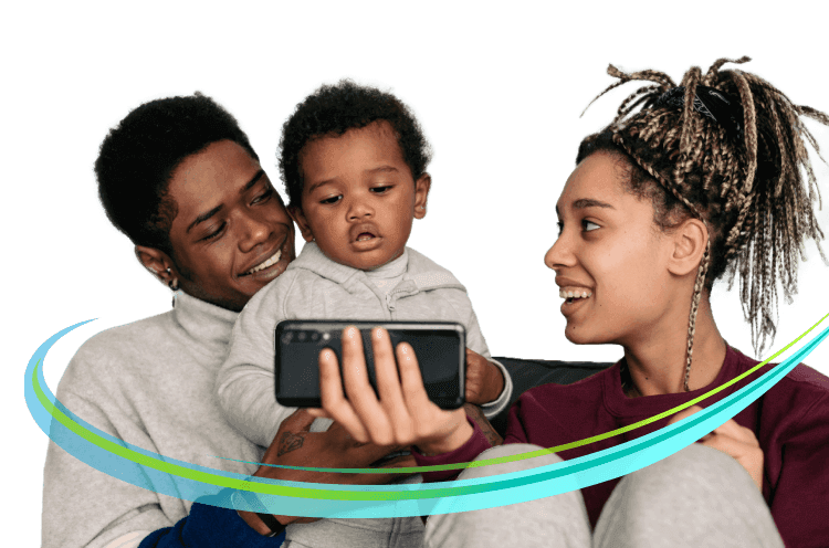 Father and mother sit with baby son on their lap. Baby looks at mobile phone mother is holding while both parents look at baby.